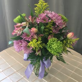 Mixed bouquet of lavender daisies, green mums, and pink blooms in a clear vase with a lavender ribbon