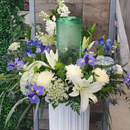 White lilies and blue-purple flowers arranged around a green glass candle holder