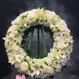 Large white and green standing wreath with roses, lilies, and daisies on a tripod stand.
