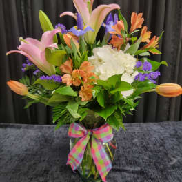 Mixed bouquet of pink lilies, orange blooms, white hydrangea and purple flowers in a glass vase with plaid bow.