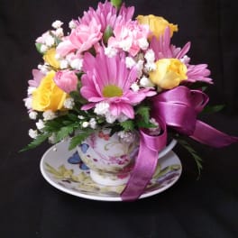 Pink and yellow flowers arranged in a teacup with a purple ribbon