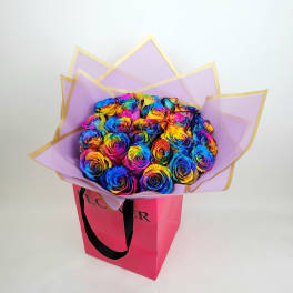 Bouquet of rainbow-colored roses in lavender wrap inside a pink gift bag