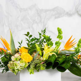 Low rectangular arrangement with yellow roses, lilies, and tropical flowers in a white container.