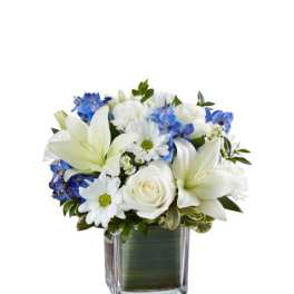 Short white and blue flower arrangement with lilies, roses, and daisies in a clear cube vase