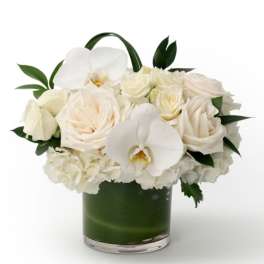Compact arrangement of white roses, hydrangeas, and orchids in a low glass vase with green lining