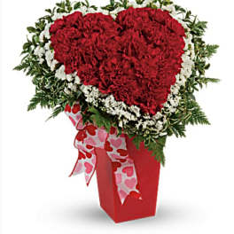 Heart-shaped red carnation arrangement in a red vase with a heart ribbon