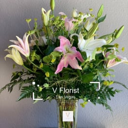 Tall arrangement of pink and white lilies with buds in a clear glass vase with ferns.