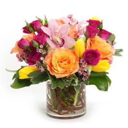 Bright arrangement of orange roses, magenta roses, yellow tulips, and pink orchids in a clear glass vase