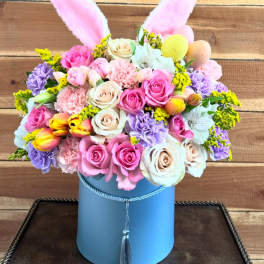 Pastel flower arrangement in a blue hatbox with pink bunny ears and decorative eggs