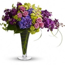 Tall arrangement of purple, blue, and green flowers in a clear trumpet vase with leaf-lined interior.