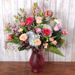 Tall mixed arrangement of pink roses, lilies, tulips, and blue hydrangeas in a red glass vase