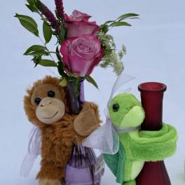 Pink roses in a purple bud vase with a monkey plush and a red vase hugged by a green turtle plush.