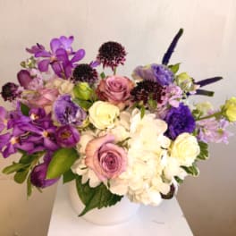 Low vase arrangement of purple orchids, lavender roses, white hydrangeas, and mixed blooms in a white pot