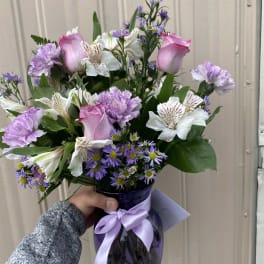 Handheld arrangement of pink roses and purple flowers in a purple glass vase with ribbon.