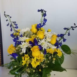 Yellow roses and white daisies in a clear glass vase with blue flowers