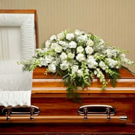 White floral casket spray on a polished wood coffin with an open white interior.