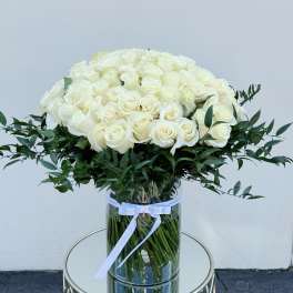 Large arrangement of cream roses with a collar of dark foliage in a tall clear cylinder vase.