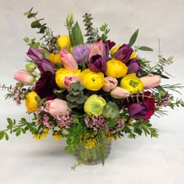 Colorful arrangement of yellow ranunculus and pink and purple tulips in a clear glass vase.