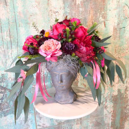 Pink and red floral arrangement in a gray bust vase with long ribbon streamers