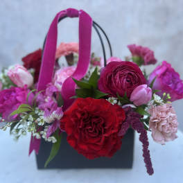 Pink and red mixed flower arrangement in a black handled box with ribbon