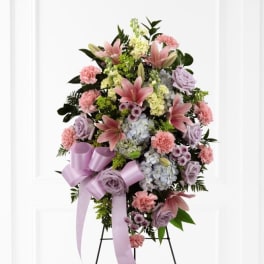 Standing spray of pink lilies, carnations, lavender roses and hydrangeas with a lavender bow on an easel.