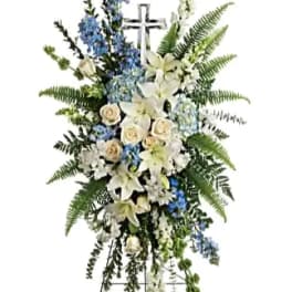 Standing funeral spray with white lilies, roses, and blue flowers on a cross easel