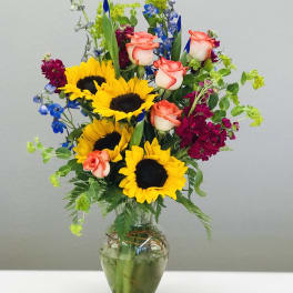Bouquet of sunflowers, coral roses, and blue flowers in a glass vase