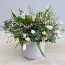 Low white ceramic pot with pink, purple, and white tulips arranged with airy greenery.