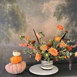 Orange floral arrangement in a striped vase beside stacked decorative pumpkins