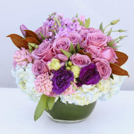 Low round arrangement of pink roses, white hydrangeas and purple flowers in a glass bowl vase