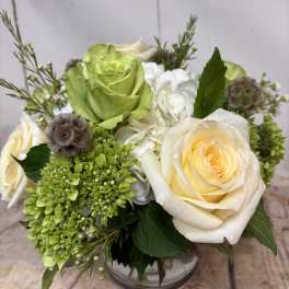 Low arrangement of cream roses, green blooms, and white hydrangeas in a clear glass vase