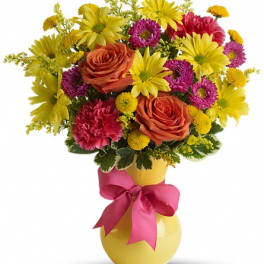 Bouquet of yellow daisies, orange roses, and pink flowers in a yellow vase with a pink ribbon