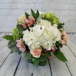 Low arrangement of white hydrangeas with peach roses and pink accents in a green glass cube vase