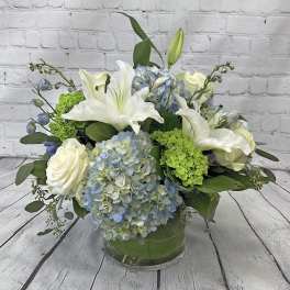 Low glass vase with white lilies, roses, blue hydrangeas, and bright green blooms in a rounded arrangement