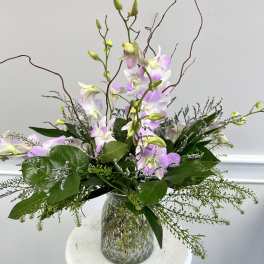 Tall arrangement of light purple dendrobium orchids with abundant greenery in a textured clear glass vase