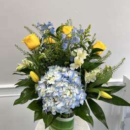 Mixed arrangement of yellow roses and tulips, blue hydrangea, and white blooms in a glass vase with green wrap