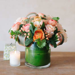 Short round arrangement of peach and cream roses in a clear glass vase with green accents