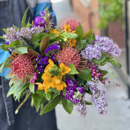 Handheld bouquet with yellow blooms, purple flowers, and pink pincushion protea