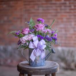 Purple and pink flower arrangement in a small metal watering can with a lavender bow