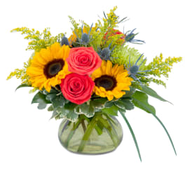 Bright arrangement of yellow sunflowers, coral roses, and blue accents in a round green glass vase