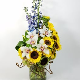 Tall bouquet of sunflowers, white orchids, and blue delphinium in a glass vase