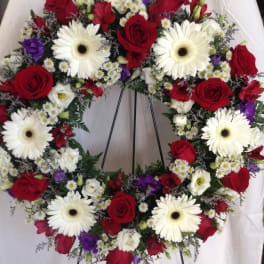 Circular wreath of red roses, white gerberas, and purple blooms on a stand