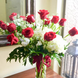 Tall arrangement of red roses and white flowers in a clear glass vase with red ribbon
