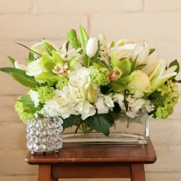 Low white and green floral arrangement in a clear glass vase beside a jeweled candle holder.
