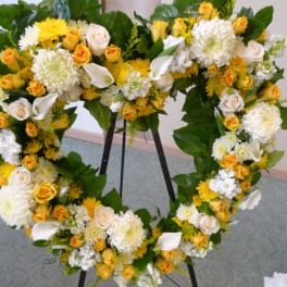Heart-shaped standing wreath of yellow and white roses, mums, and calla lilies on a metal easel.