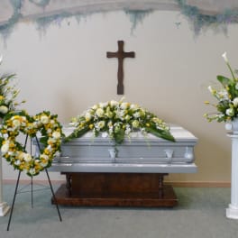 Funeral casket with white and yellow floral sprays, heart wreath, and pedestal arrangements.