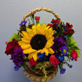 Basket arrangement with a large sunflower and mixed red, purple, and blue flowers