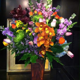 Tall mixed arrangement with orange orchids, purple flowers, and tulips in a brown glass vase
