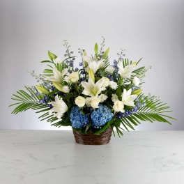 White lilies and roses with blue hydrangeas in a fan-shaped basket with palm fronds.