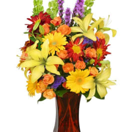 Tall mixed bouquet of yellow lilies, gerbera daisies and orange roses in an amber glass vase